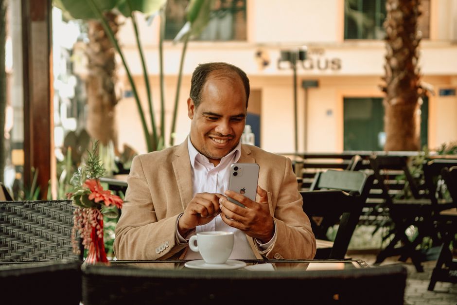 A man in a white shirt smiles while using his smartphone at an outdoor café.